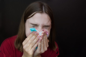 Sick young girl with allergies blows her nose in a napkin. Dark background. Copyspace.