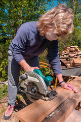 masked woman using a circular saw cuts a wooden board on the street in the early summer morning.