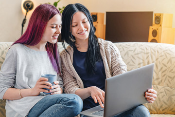 Young happy girls sitting on sofa at home drinking coffee while using laptop