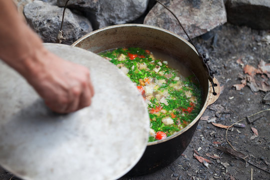 Freshly Cooked Hot Soup In Old Cauldron On Camp Fire And Man Hand With Pot Cover