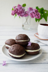 Several Whoopie Pies on a white plate on a white textured wooden background. Vertical orientation with lilac blossom.