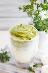 Dalgona matcha coffee or tea - green iced coffee drink with milk on white table with plants, sunny light. New trend drink in social media. Morning healthy reverse latte. Selective focus, copy space.