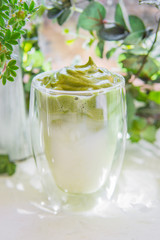 Dalgona matcha coffee or tea - green iced coffee drink with milk on white table with plants, sunny light. New trend drink in social media. Morning healthy reverse latte. Selective focus, copy space.