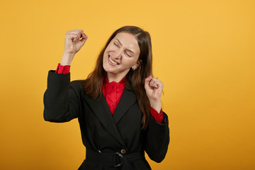 Young Attractive Brunette Woman In Black Stylish Suit, Red Shirt On Yellow Background, Happy Girl Smiles, Clenched Her Hands Into Fists, Raised Them Up. The Concept Of Kind, Cheerful People
