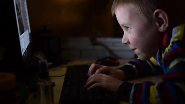 Little boy is typing on the keyboard. The child indulges, plays at the computer in a dark room.
