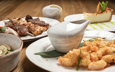 composition of shish kebab, fried prawns, and cheesecake dessert on white plates and a wooden table