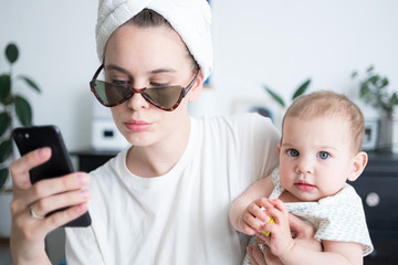 Woman with towel around head in sunglasses holding little baby and chatting, taking photos in mobile phone. motherhood family lifestyle concept. morning routine