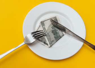 A hundred-dollar bill lies on a white plate near a fork and a knife on a yellow cotton tablecloth....