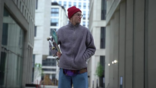 Young readhead hispter keeps skateboard on the soulder and walks the street looking to the camera. Outdoor. Cloudy. Portrait