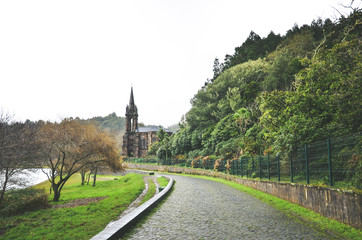Cobbled path by the lake surrounded by green trees. Chapel of Nossa Senhora das Vitorias in Furnas, Azores Islands, Portugal in the background. Overcast, rainy day, white sky. Horizontal photo.