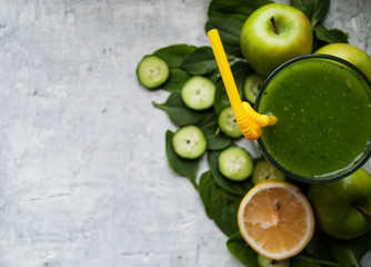 Glass with green smoothie, composition: spinach, cucumber, apple, lemon, sorrel. Detox drink and its components beautifully laid out on a gray background. Bright yellow cocktail straw.
