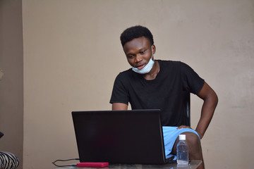 a black boy attending online class with his laptop and wearing medical face mask. hand sanitizer on the table