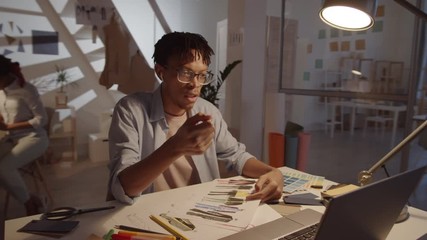 360-degree dolly shot of African American male designer in wireless earphones discussing color palette and fashion illustrations with colleague via online video call on laptop at office desk - Powered by Adobe