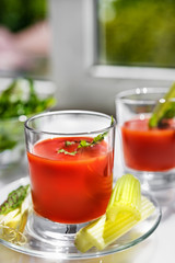 Two glasses of tomato juice with parsley and celery decorations, stand near the window, morning sunlight shines, shallow depth of field, selective focus. Natural drinks concept.