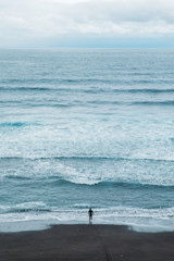 Fototapeta na wymiar The surfer is heading towards the high waves of the ocean. The Atlantic ocean and the black beach. Vertical landscape from above.