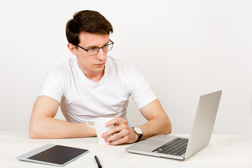 A handsome man with a light smile wearing a T-shirt and glasses sits against a white wall at his desk and work with a laptop. The concept of remote quarantine education and communication