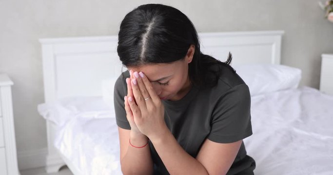 Hopeful young african american woman sitting on bed with folded hands, saying morning worship. Faithful 30s mixed race lady praying God, asking for help or waiting for miracle alone in bedroom.