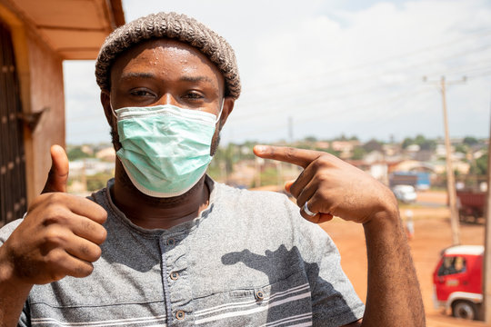 portrait of a young african man wearing a face mask outside to protect against the coronavirus, doing a thumbs up gesture and pointing to his face mask
