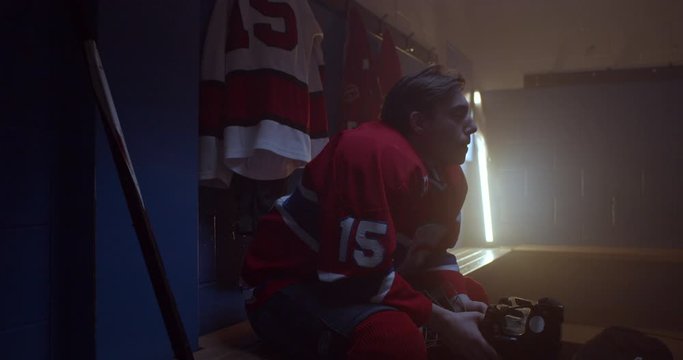 Hockey player sits in dramatically lit changer oom holding helmet with determination and focus in his eyes determined to win a crucial game in series. Puts on hockey equipment, 4K