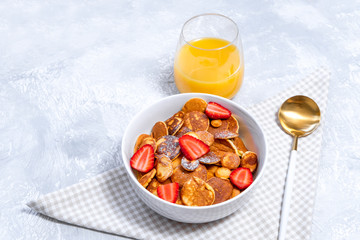 Tiny pancakes cereal with strawberry and orange juice glass on concrete backdrop. Trendy breakfast, american food, space for text