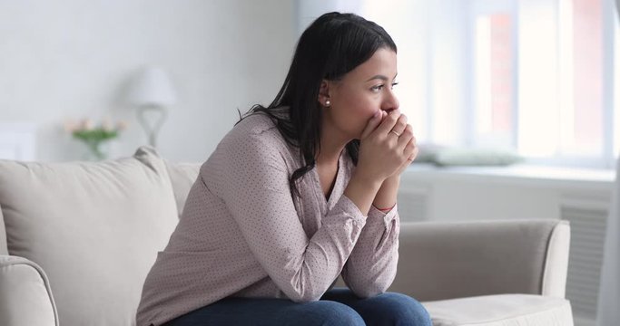 Frustrated african ethnicity young woman sitting on couch, thinking of personal problems alone at home. Depressed unhappy mixed race lady suffering from mental stress, feeling unsure of decision.