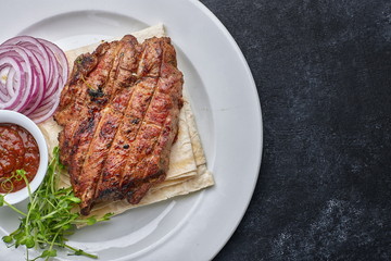 Grilled meat steak with onions, sauce and microgreen, on pita bread, on a white plate, against a dark background. space for text. top view