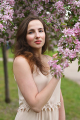 A young beautiful woman with a slight smile touches a blooming branch of an Apple tree