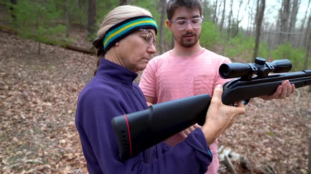 Teen son teaching his mother how to hold and fire a pellet rifle outside.