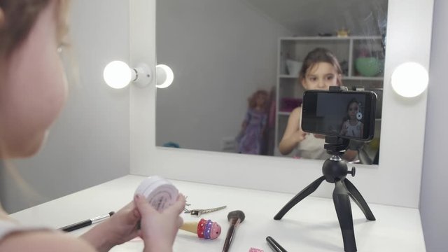 Kid makes selfie using phone and tripod. Brunette girl takes photos at home. Girl is learning how to be photographer.