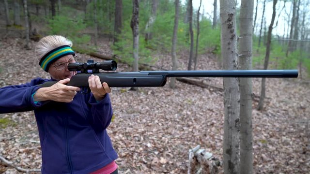 Mature woman aims pellet rifle and son tries to help her steady it then she fires it and gets angry holding her ear.