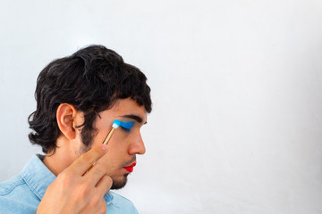 Non-binary bearded young Hispanic man on a white background, putting on extravagant makeup with a brush, red lips and blue eye shadows