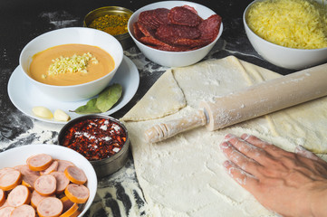 Pizza ingredients. hand rolls out the dough. rolling pin and flour. sliced salami and sausages. grated cheese and pizza sauce