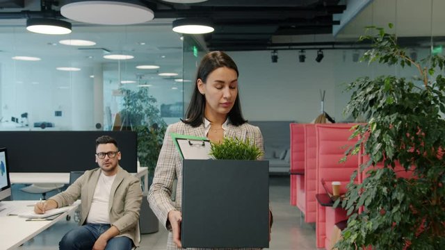 Sad young woman fired employee is leaving work with box of stuff walking in open space office while coworkers watching her with unhappy faces.