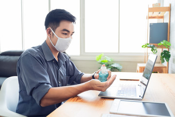 Asian businessman work from home self isolation in quarantine, stay safe with gel sanitizer cleaning hands and wearing facemask from coronavirus covid-19 infection, office room using computer laptop
