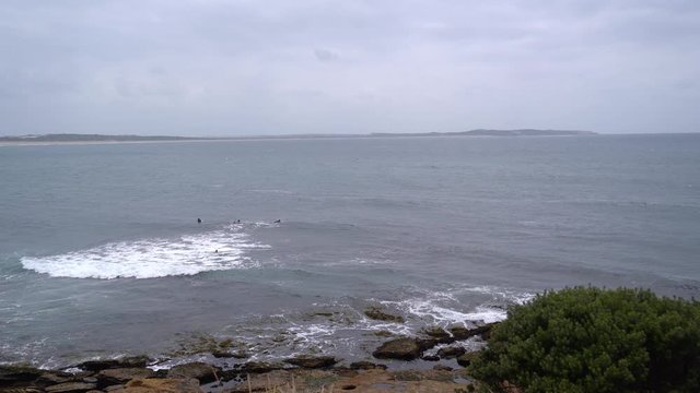 A view on an ocean bay framed by green shrubs and rocky beach with surfers waiting for waves in the ocean. Ocean horizon in distance under a greay gloomy sky.