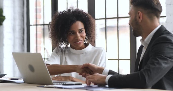 Smiling young african ethnicity businesswoman making agreement with male partner, shaking hands, establishing partnership. Confident businessman signing contract document deal with biracial client.