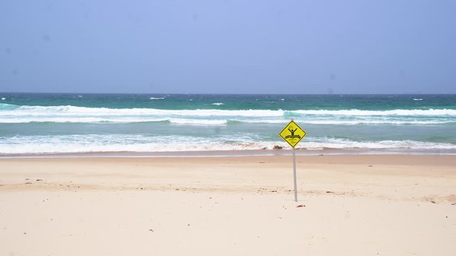 A warning sign of dangerous rip current on a lonely white sand with beach no people around. Blue waves are hitting the sand in a strong wind under a cloudless sunny blue sky.