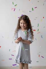 Charming curly brunette girl in a beautiful white dress with her hair is having fun, clapping her hands, playing with multi-colored falling confetti. Happy childhood. A little party .