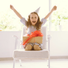 Happy delighted little girl sitting at home on the armchair on her birthday. The festive cap is dressed on the head. Delight and joy on the face of the child.