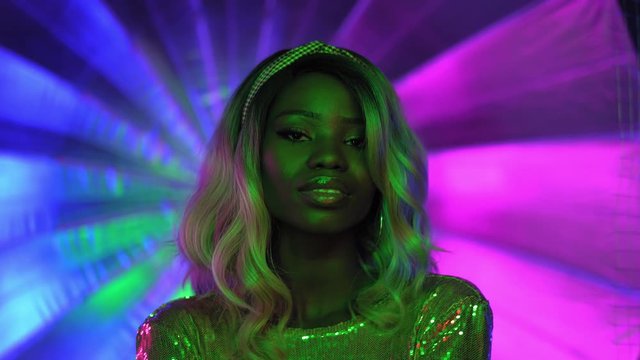A blonde African-American woman with green lighting on her face stands on a beautiful colored neon-lit background in neon light, dressed in a silver sequin dress. Neon concept. Prores 422. 