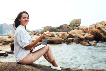 Fototapeta na wymiar Young successful, smiling girl working using a laptop, sitting on a rocky seashore. traveling, blogger, freelancer, content plan, work online