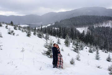 Fototapeta na wymiar The bride and groom stand against the backdrop of the mountains and cuddle under a rug to keep warm. Winter wedding