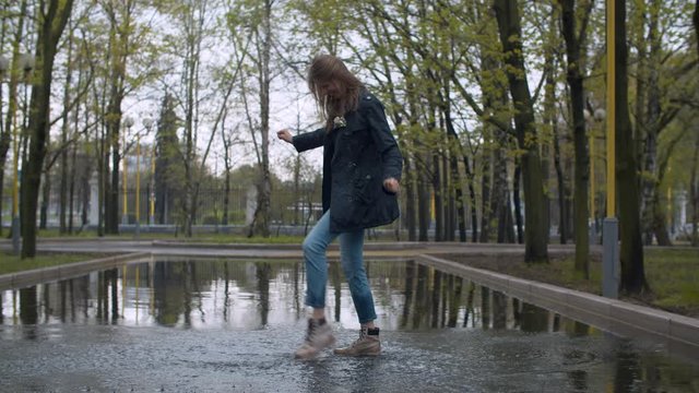 Happy woman jumping in a deep puddle in large splashes. Trees are reflected in a puddle. Rainy day in the park. Real people