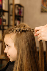 Naklejka premium Process of braiding the master weaves braids on her head blond little girl in beauty salon close up. Professional hair care and creating hairstyles.