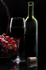 A wine bottle and a cup of wine with a basket of grapes on the side in low key light or dark food...