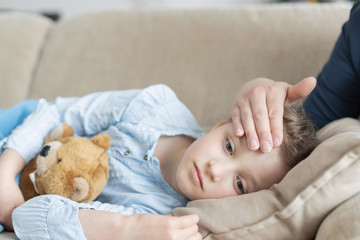 A sick and sad cute girl lies on a sofa with a soft toy and her mother checks the heat or temperature on her forehead. sick children, flu