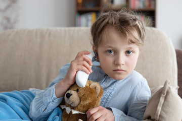 Caucasian cute sad girl measures the temperature in her ear with a thermometer at home. Sick child at home, fever, flu