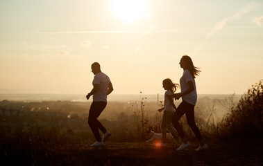 Fototapeta na wymiar Young and happy family - man, woman and girl are jogging with their dog outside the city, on the village road at sunset, side view, copy space