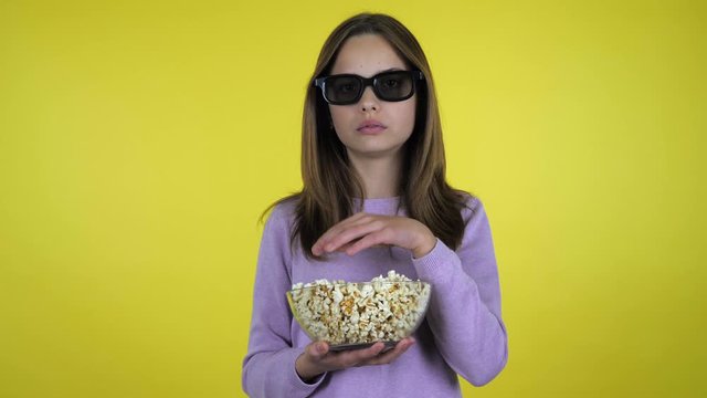 Teenager girl in a pink sweater watches movie in black 3D glasses, eats popcorn from glass bowl and is unhappy with the taste on yellow background with copy space. Place for text or product
