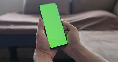 Closeup young man sit on a couch and watch something on a smartphone with green screen